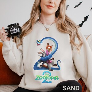 May include: Sand-colored sweatshirt featuring a graphic from Disney's Zootopia 2. The design includes characters Nick Wilde, Judy Hopps, and a blue snake. The sweatshirt has a crew neck and long sleeves. The word "SAND" is printed at the bottom.
