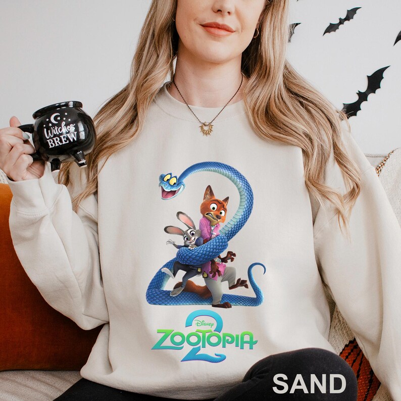 May include: Sand-colored sweatshirt featuring a graphic from Disney's Zootopia 2. The design includes characters Nick Wilde, Judy Hopps, and a blue snake. The sweatshirt has a crew neck and long sleeves. The word "SAND" is printed at the bottom.