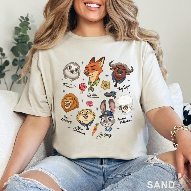 Puede incluir: Una camiseta color arena con un estampado gr&aacute;fico de los personajes de Zootopia, incluyendo a Nick Wilde, Judy Hopps, Flash, Chief Bogo, Mayor Lionheart, Officer Clawhauser y Assistant Bellwether, junto a iconos como un donut y una zanahoria.