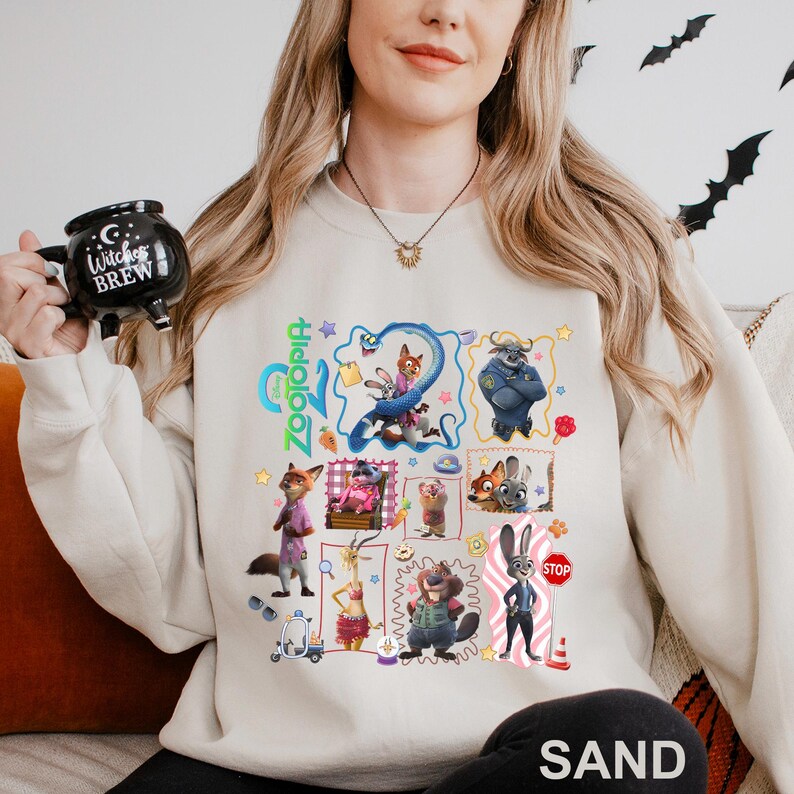 May include: Sand-colored sweatshirt featuring a colorful graphic of characters from the animated film "Zootopia." The design includes various character illustrations and the film's title. The word "SAND" is printed at the bottom.
