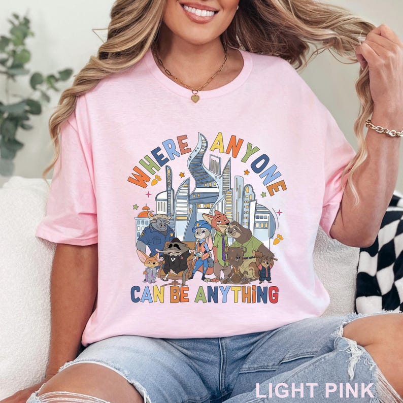 May include: Light pink t-shirt with a colorful graphic featuring cartoon characters and the text "WHERE ANYONE CAN BE ANYTHING". The design includes a cityscape and various animal figures. The shirt is made of a soft material.