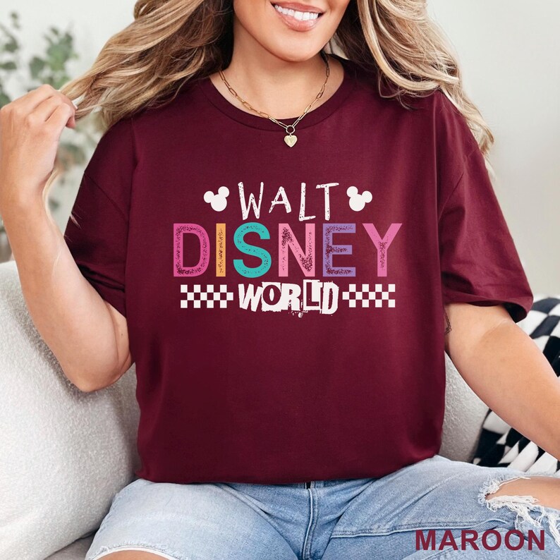 Pu&ograve; includere: Una t-shirt a maniche corte color bordeaux con la scritta WALT DISNEY WORLD in lettere colorate e testurizzate. Il design include due piccole sagome bianche della testa di Topolino sopra la parola WALT e un motivo a scacchi bianco sotto WORLD.