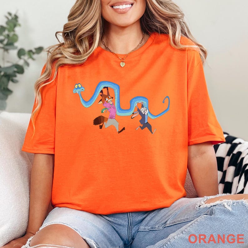 May include: A bright orange short-sleeved t-shirt featuring a graphic print of a long blue snake winding around a cartoon fox wearing a pink shirt and a cartoon rabbit wearing a blue jacket. The characters appear to be running in a forward motion.
