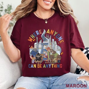 May include: Maroon t-shirt featuring a colorful graphic of various cartoon characters in front of a cityscape. The text on the shirt reads "WHERE ANYONE CAN BE ANYTHING". The shirt is made of a soft material.