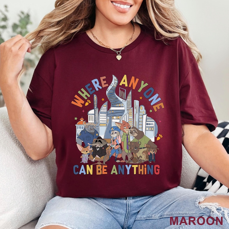 May include: Maroon t-shirt featuring a colorful graphic of various cartoon characters in front of a cityscape. The text on the shirt reads "WHERE ANYONE CAN BE ANYTHING". The shirt is made of a soft material.
