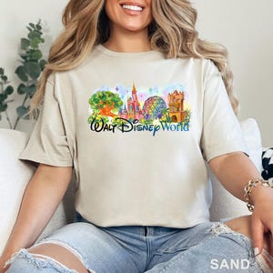 May include: A sand-colored short-sleeve t-shirt featuring a colorful graphic print of the Walt Disney World theme park landmarks, including the castle, the geodesic sphere, and the tree of life, with the text Walt Disney World in blue script.