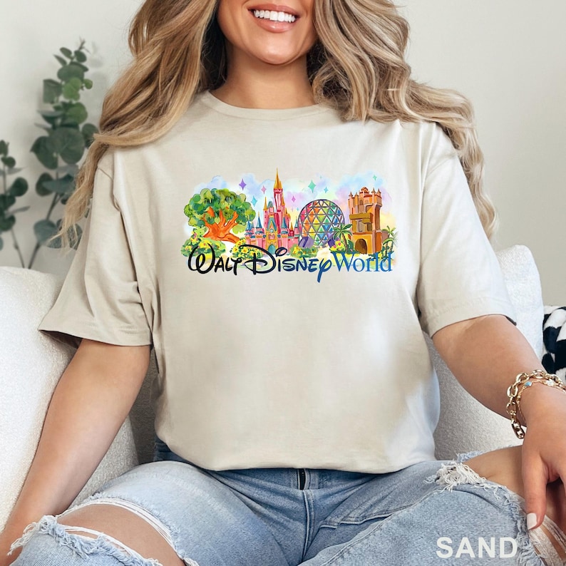 May include: A sand-colored short-sleeve t-shirt featuring a colorful graphic print of the Walt Disney World theme park landmarks, including the castle, the geodesic sphere, and the tree of life, with the text Walt Disney World in blue script.