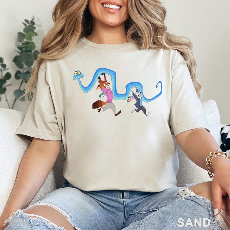 May include: A sand-colored t-shirt featuring a graphic print of a long blue snake being held up by a cartoon fox wearing a pink shirt and a cartoon rabbit wearing a blue uniform. The characters are depicted in a dynamic, running pose together.