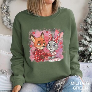 May include: Military green sweatshirt featuring a cartoon fox and bunny holding a heart-shaped gift. The fox wears a red scarf and sweater with white hearts. The bunny has a red bow and dress with heart patterns. Pink background with hearts.