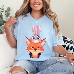 May include: Pale blue t-shirt with a cartoon illustration of Judy Hopps and Nick Wilde from Zootropolis. The text "LIGHT BLUE" is printed at the bottom. The design features a grey rabbit and an orange fox.