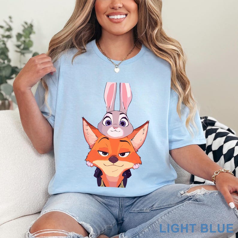 May include: Pale blue t-shirt with a cartoon illustration of Judy Hopps and Nick Wilde from Zootropolis. The text "LIGHT BLUE" is printed at the bottom. The design features a grey rabbit and an orange fox.
