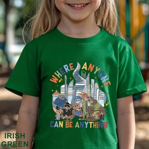 May include: Irish green t-shirt with a colorful graphic featuring cartoon characters and the text "WHERE ANYONE CAN BE ANYTHING". The design includes a cityscape and various animal figures.