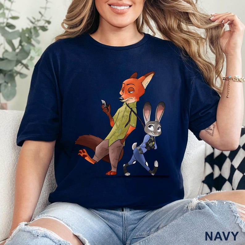 May include: Navy blue t-shirt featuring a cartoon illustration of Nick Wilde the fox and Judy Hopps the rabbit from Zootopia. Nick is holding a phone, and Judy is in a police uniform. The word "NAVY" is printed at the bottom.