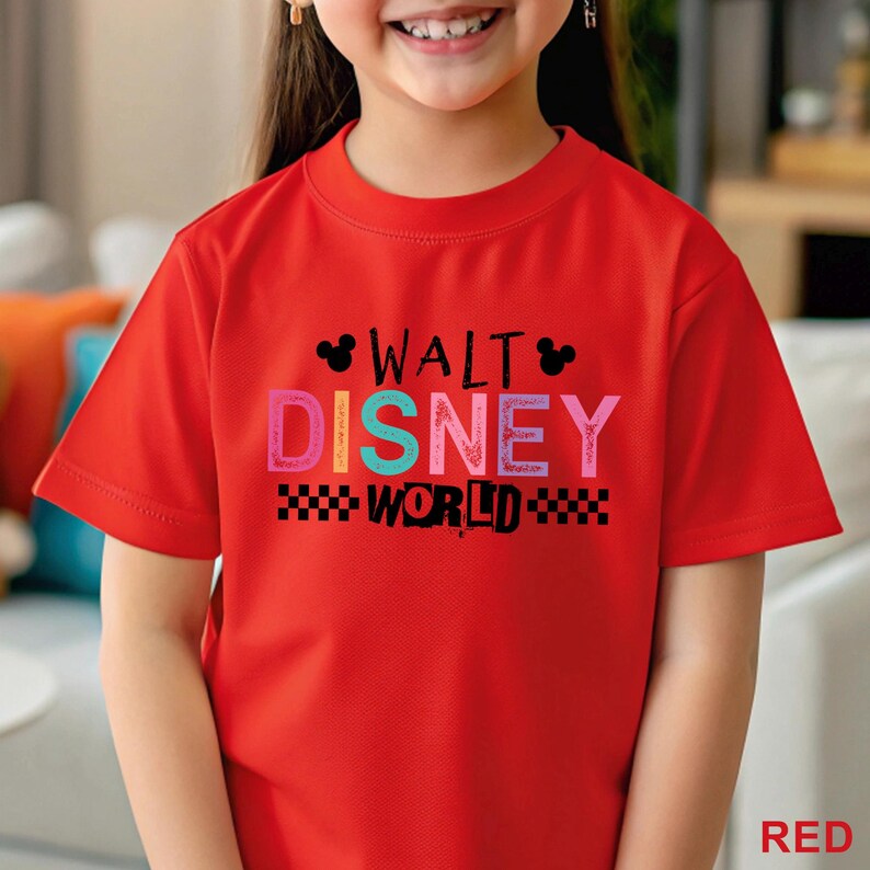 Pu&ograve; includere: Una t-shirt rossa a maniche corte con la scritta Walt Disney World in un carattere multicolore. Il design include due piccole sagome nere delle orecchie di Topolino e motivi a scacchi neri ai lati della parola World sul davanti.