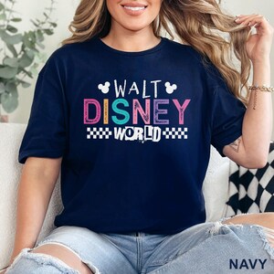 Pu&ograve; includere: Una t-shirt blu navy a maniche corte con la scritta WALT DISNEY WORLD in un carattere multicolore e dall'aspetto vissuto. Il design include due sagome bianche delle orecchie di Topolino e un motivo a scacchi bianco sotto la parola WORLD.