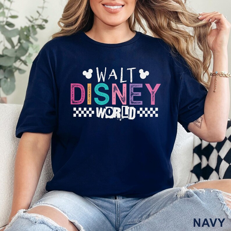 Pu&ograve; includere: Una t-shirt blu navy a maniche corte con la scritta WALT DISNEY WORLD in un carattere multicolore e dall'aspetto vissuto. Il design include due sagome bianche delle orecchie di Topolino e un motivo a scacchi bianco sotto la parola WORLD.