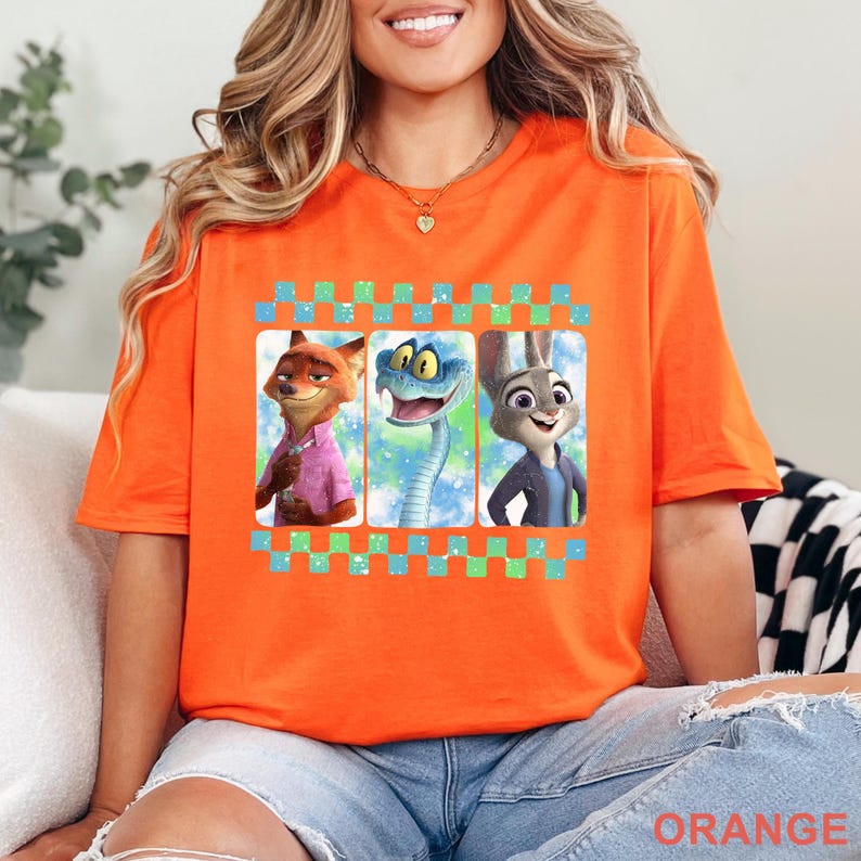 May include: Orange t-shirt featuring three cartoon characters in a checkered frame. The characters include a fox in a pink shirt, a blue snake, and a rabbit in a blue jacket. The word "ORANGE" is printed at the bottom.
