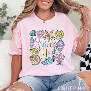 May include: Light pink t-shirt featuring a colorful seashell design with the text "Shell Yeah". The design includes various seashells and a starfish in shades of blue, yellow, and pink. The shirt is made of soft material.
