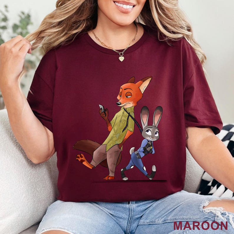 May include: Maroon t-shirt featuring a cartoon illustration of Nick Wilde and Judy Hopps from Zootopia. Nick is depicted holding a phone, while Judy is in a police uniform. The word "MAROON" is printed at the bottom.