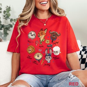 Puede incluir: Una camiseta de color rojo brillante con un estampado gr&aacute;fico colorido de varios personajes animales animados de Zootopia, incluidos Nick Wilde, Judy Hopps, Flash, Chief Bogo, Mayor Lionheart, Officer Clawhauser y Assistant Bellwether.