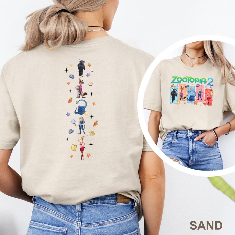 May include: Sand-colored t-shirt featuring a colorful graphic of characters from Zootopia 2. The back of the shirt has a vertical design with various character illustrations and decorative elements. The shirt is made of a soft material.