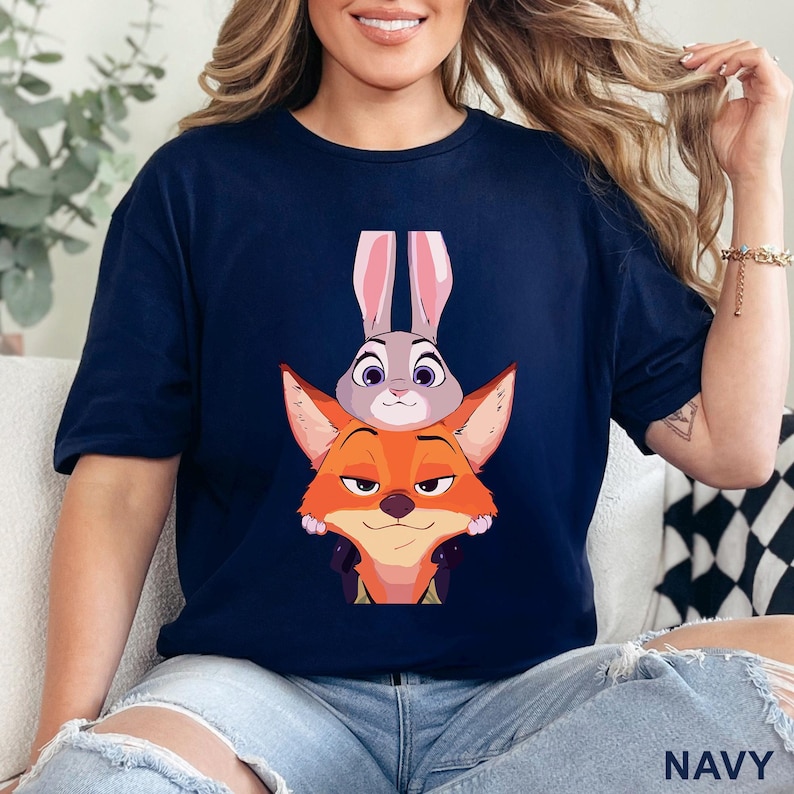 May include: Navy blue t-shirt with a cartoon illustration of Judy Hopps the rabbit and Nick Wilde the fox from Zootopia. The graphic shows the characters stacked on top of each other. The word "NAVY" is printed at the bottom of the shirt.