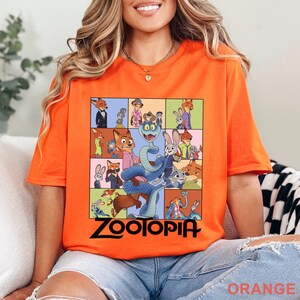 May include: A bright orange short-sleeve t-shirt featuring a grid of various cartoon characters from the movie Zootopia, including a large blue snake in the centre. The word Zootopia is printed in bold black lettering at the bottom of the graphic.