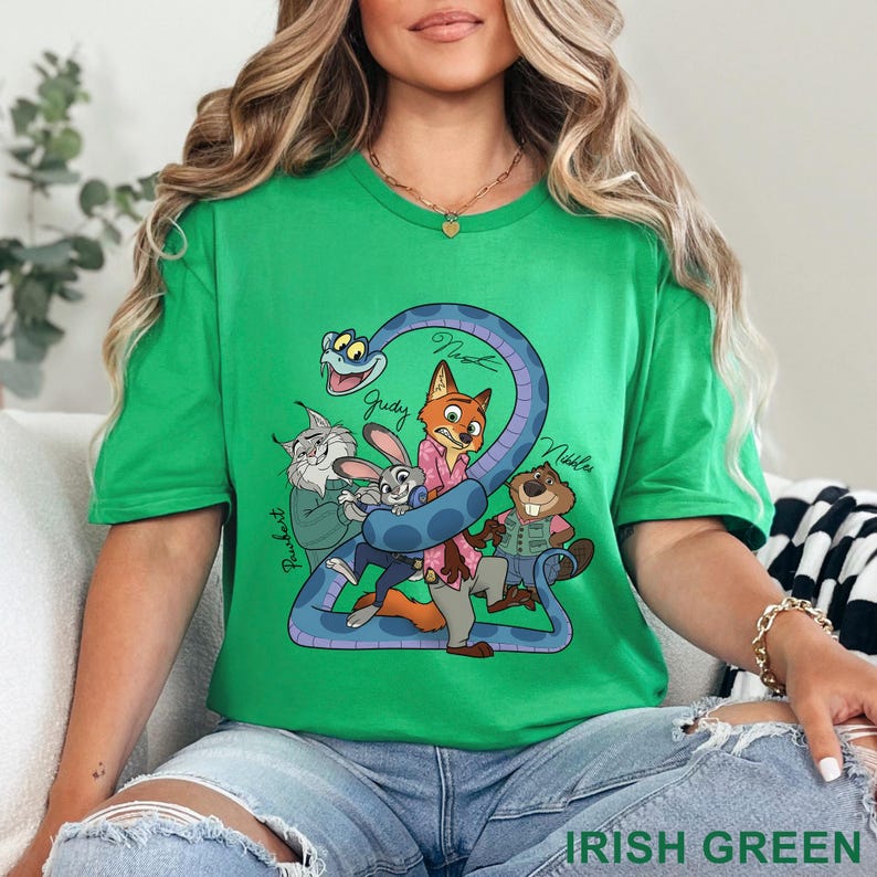 Puede incluir: Una camiseta de manga corta de color verde brillante con un estampado gr&aacute;fico colorido de personajes animados, incluyendo una serpiente, un zorro, un conejo, un lince y un castor. El texto "Judy", "Nick", "Nibbles" y "Pawbert" aparece impreso cerca.