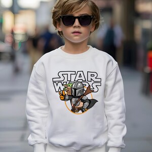 May include: White sweatshirt with a Star Wars graphic. The graphic features the text "STAR WARS" above a cartoon illustration of The Mandalorian and Grogu. The Mandalorian is holding a weapon, and Grogu is in a carrier.