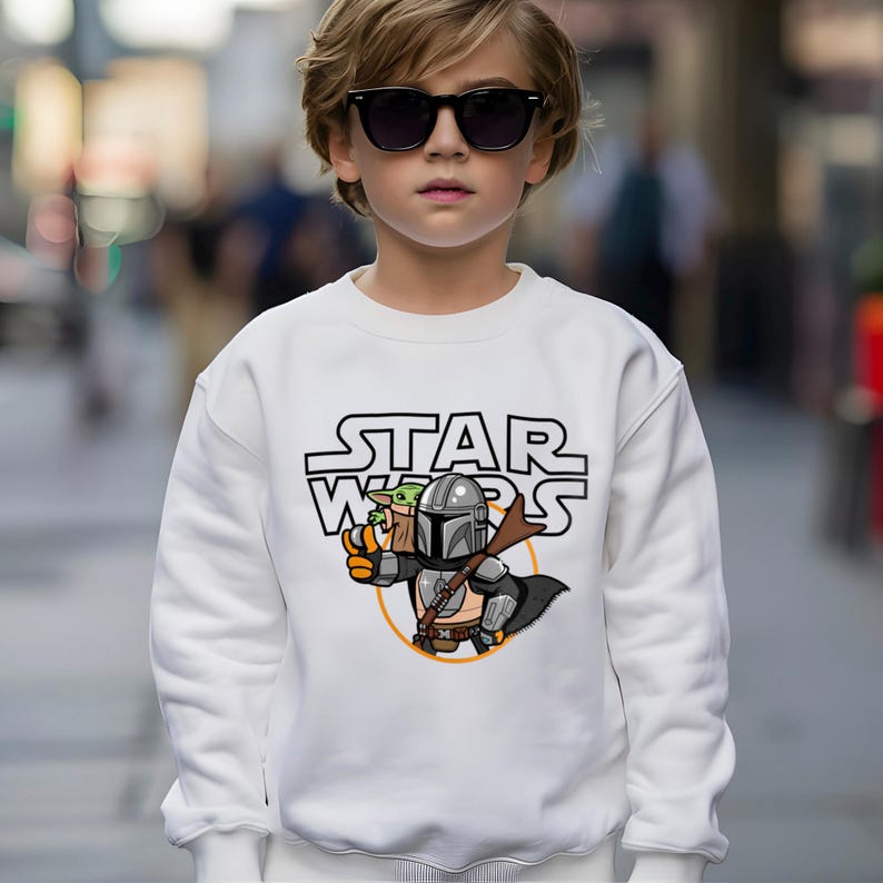 May include: White sweatshirt with a Star Wars graphic. The graphic features the text "STAR WARS" above a cartoon illustration of The Mandalorian and Grogu. The Mandalorian is holding a weapon, and Grogu is in a carrier.
