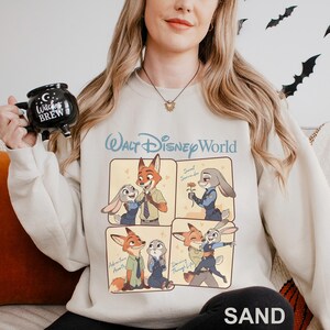 Puede incluir: Sudadera de color arena con un dise&ntilde;o de dibujos animados con personajes de Zootopia. El dise&ntilde;o incluye el texto "Walt Disney World" y cuatro paneles que representan a los personajes. La palabra "SAND" est&aacute; impresa en la parte inferior.