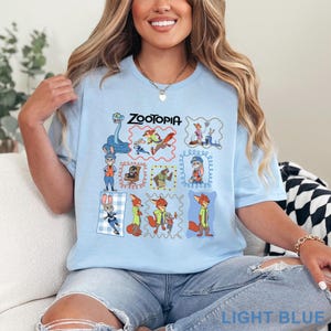 K&ouml;nnte beinhalten: Ein hellblaues T-Shirt mit einem Grafikdruck verschiedener Zootopia-Figuren, darunter Judy Hopps und Nick Wilde. Das Design enth&auml;lt mehrere gerahmte Illustrationen der Figuren in verschiedenen Posen und das Wort Zootopia am oberen Rand.