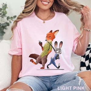 May include: Light pink t-shirt featuring a cartoon illustration of a fox and a rabbit. The fox is holding a phone and the rabbit is walking. The text "LIGHT PINK" is at the bottom of the shirt.
