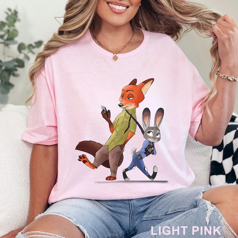 May include: Light pink t-shirt featuring a cartoon illustration of a fox and a rabbit. The fox is holding a phone and the rabbit is walking. The text "LIGHT PINK" is at the bottom of the shirt.