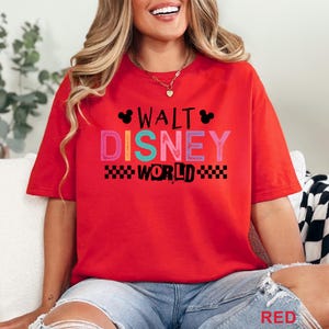 Pu&ograve; includere: Una t-shirt rossa a maniche corte con la scritta WALT DISNEY WORLD in un carattere a blocchi multicolore. Il design include sagome nere delle orecchie di Topolino e un motivo a scacchi bianco e nero sotto la parola WORLD.