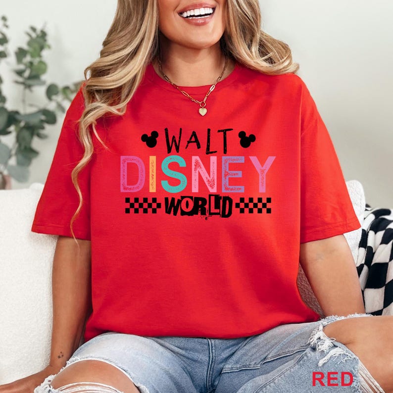 Pu&ograve; includere: Una t-shirt rossa a maniche corte con la scritta WALT DISNEY WORLD in un carattere a blocchi multicolore. Il design include sagome nere delle orecchie di Topolino e un motivo a scacchi bianco e nero sotto la parola WORLD.