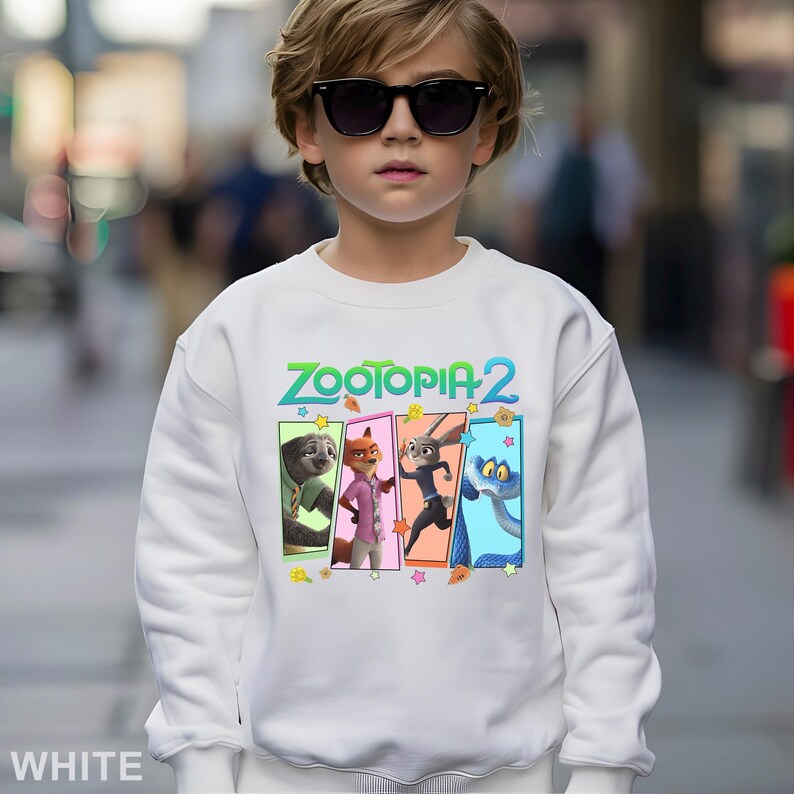 May include: White sweatshirt featuring the text "Zootopia 2" in green and blue. The design includes cartoon characters in colorful rectangular frames. The sweatshirt is worn by a person wearing sunglasses.