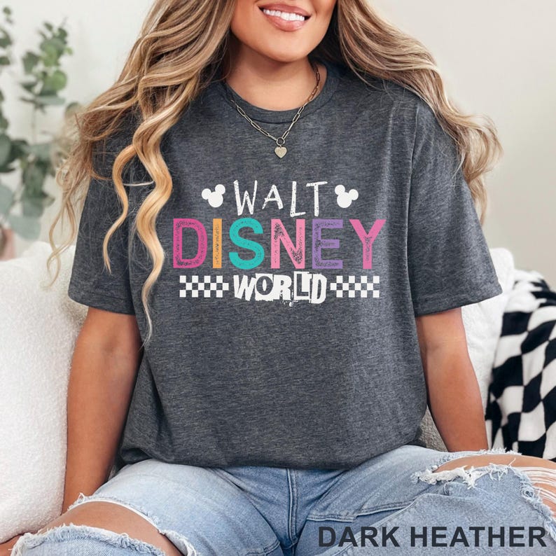 Pu&ograve; includere: Una t-shirt a maniche corte grigio melange scuro con la scritta bianca WALT, la scritta colorata DISNEY e la scritta bianca WORLD con un motivo a scacchi. Il design include due piccole sagome bianche di orecchie di Topolino sopra il testo.
