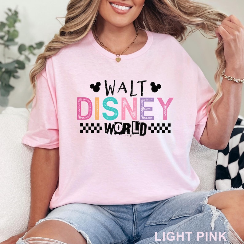 Pu&ograve; includere: Una t-shirt a maniche corte rosa chiaro con la scritta WALT DISNEY WORLD in un carattere multicolore dall'aspetto vissuto. Il design include due sagome nere di orecchie di Topolino e motivi a scacchi bianchi e neri ai lati della parola WORLD.