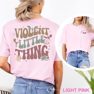 May include: Light pink t-shirt with the text "VIOLENT LITTLE THING" in brown, green, and pink lettering. The design includes illustrations of bats, a book, and other fantasy elements. The shirt is a light pink color.