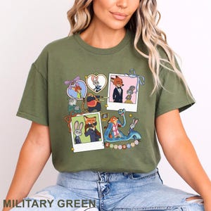 May include: An olive green t-shirt featuring a graphic print of Zootopia characters. The design includes multiple framed images, polaroid-style photos, and illustrations of various animals from the movie, with the word Zootopia at the bottom.