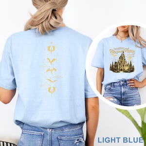 May include: Light blue t-shirt featuring a gold dragon and cloud design on the back. The front displays a castle graphic with the text "Riverrun House". The shirt is light blue.