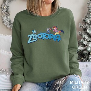 Peut inclure: Sweat-shirt vert militaire avec le mot "Zootopia" en bleu et des personnages de dessins anim&eacute;s. Le sweat-shirt est de style col rond. Le texte "MILITARY GREEN" est imprim&eacute; en blanc en bas &agrave; droite.