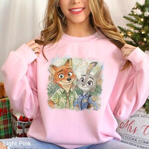 Puede incluir: Sudadera rosa claro con un estampado colorido de Nick Wilde y Judy Hopps de Zootopia. La sudadera tiene cuello redondo y mangas largas. El texto "Light Pink" es visible en la parte inferior izquierda.