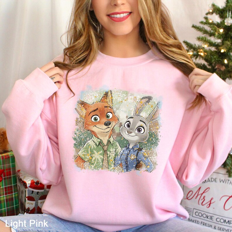 Puede incluir: Sudadera rosa claro con un estampado colorido de Nick Wilde y Judy Hopps de Zootopia. La sudadera tiene cuello redondo y mangas largas. El texto "Light Pink" es visible en la parte inferior izquierda.