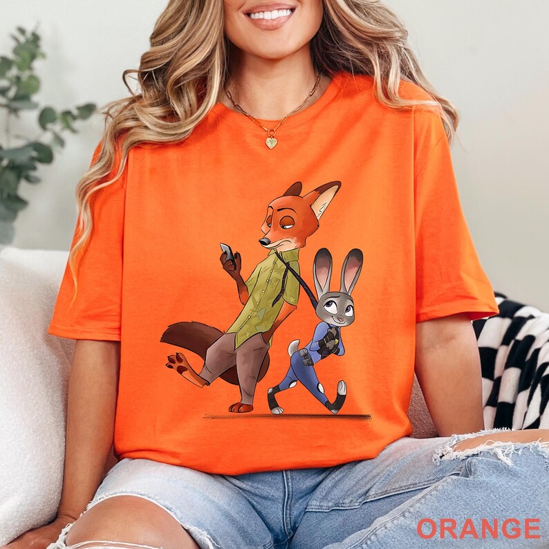 May include: Orange t-shirt featuring cartoon characters Nick Wilde and Judy Hopps from Zootopia. Nick, a fox, is holding a phone, while Judy, a rabbit, walks beside him. The word "ORANGE" is printed at the bottom of the shirt.