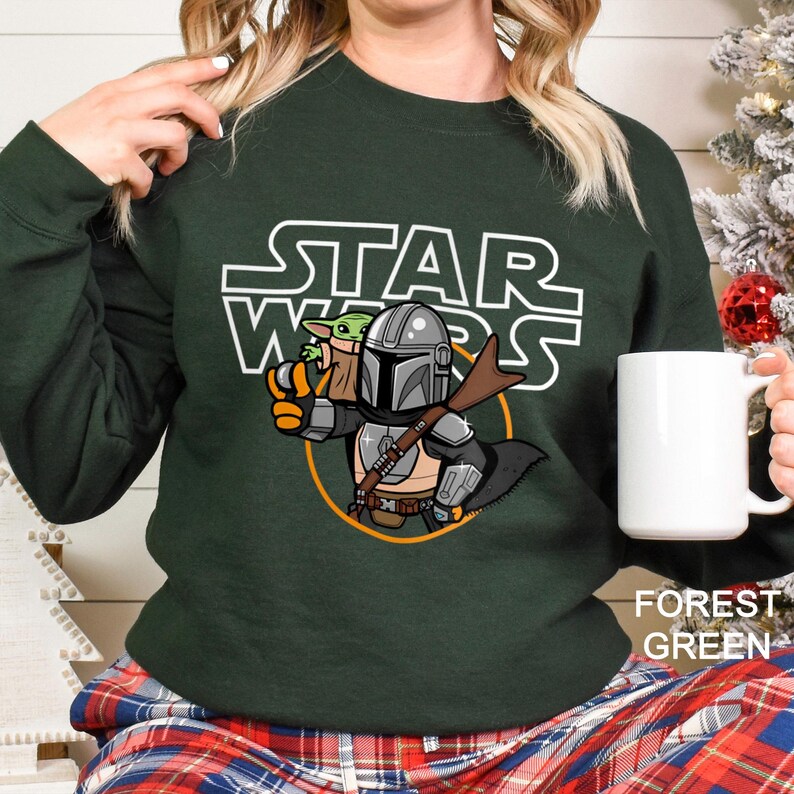 May include: Forest green sweatshirt featuring a Star Wars graphic. The design includes the Star Wars logo, the Mandalorian, and Grogu. The sweatshirt has a crew neck and long sleeves. The text "FOREST GREEN" is printed on the bottom right.