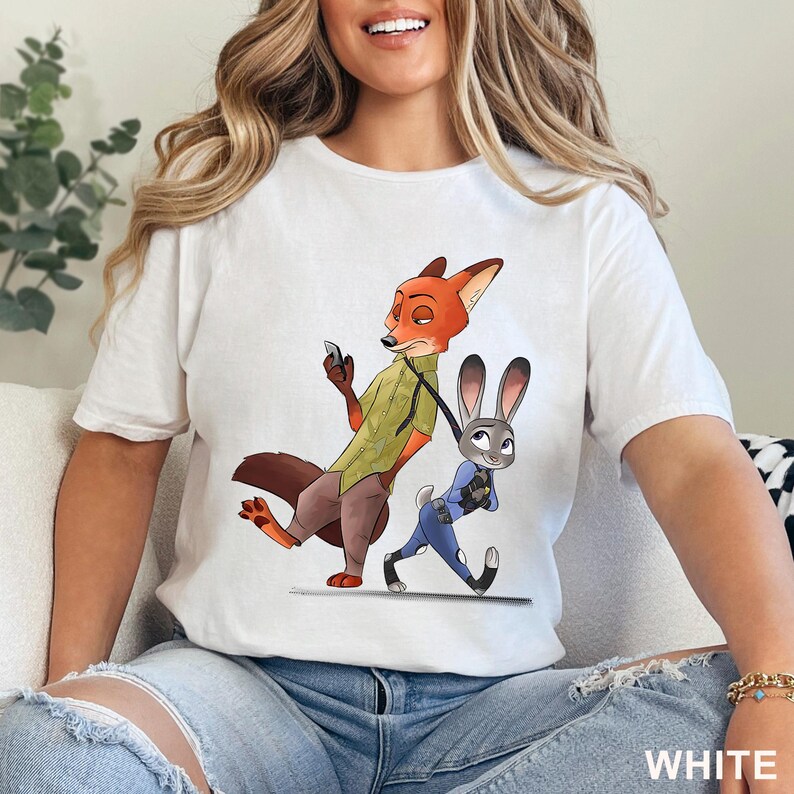 May include: White t-shirt featuring a cartoon illustration of Nick Wilde and Judy Hopps from Zootopia. Nick, a fox, is holding a phone, while Judy, a rabbit, walks beside him. The word "WHITE" is printed at the bottom.