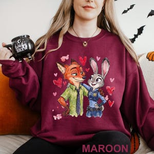 May include: Maroon sweatshirt with a cartoon illustration of Nick Wilde and Judy Hopps from Zootopia, surrounded by hearts. The word "MAROON" is printed at the bottom. A black mug with the text "Witches Brew" is held in the upper left corner.