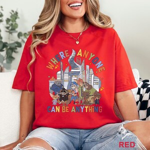 May include: Red t-shirt with a colorful graphic featuring cartoon characters and the text "WHERE ANYONE CAN BE ANYTHING." The design includes a cityscape and various animal figures. The shirt is a casual, oversized fit.